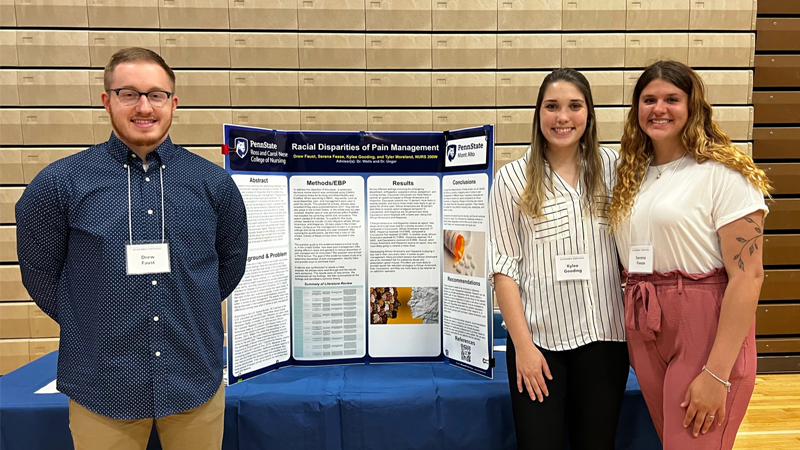 One male and two female students standing by a tri-panel research poster on a table. 